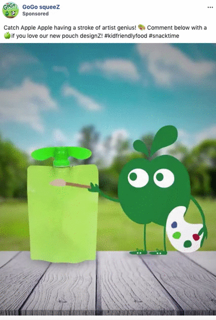 An animated green apple character with a paint palette points a brush at a green GoGo squeeZ pouch on a picnic table with a blurred outdoor background.