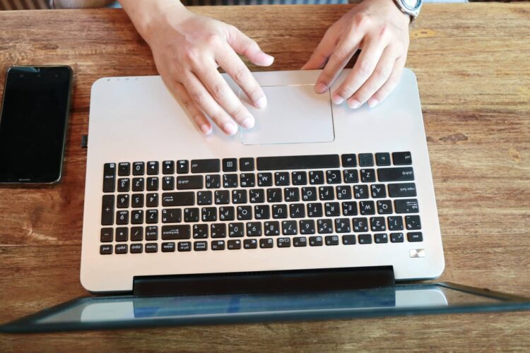 Person using a laptop on a wooden table with a smartphone placed to the left of the laptop.