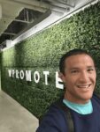 A person smiling in front of a green hedge wall with large white letters spelling "WPROMOTE" in an indoor hallway.