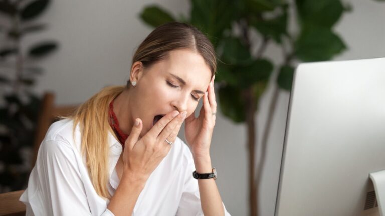 A woman sitting at a desk in front of a computer yawns and rests her head on one hand, appearing tired or fatigued.