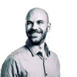 A man with a shaved head and beard smiles while looking to the side. He is wearing a button-down shirt and is set against a plain white background.