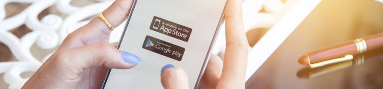 A person with painted nails holds a smartphone displaying App Store and Google Play download buttons; a pen and papers are blurred in the background.