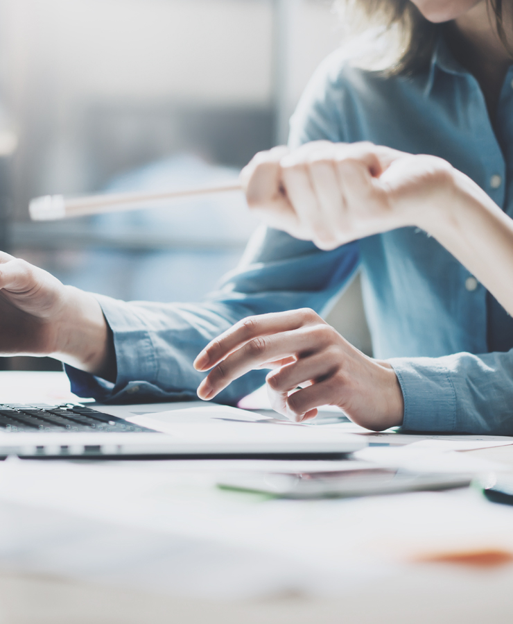 Two people sit at a desk discussing something on a laptop screen, with one person holding and pointing with a pencil.