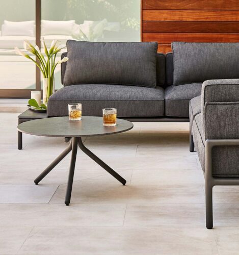 A modern outdoor patio with a gray sofa, a matching armchair, a round black coffee table, and two glasses with drinks on the table. White lilies are in a vase in the background.