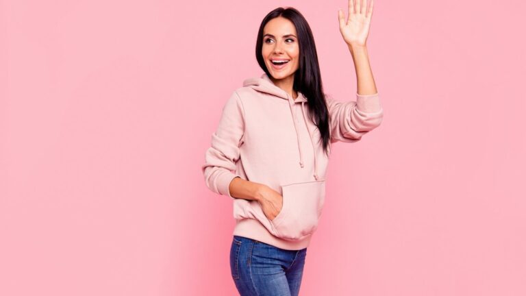 Woman with long dark hair wearing a light pink hoodie and blue jeans stands against a pink background, smiling and raising her right hand in a wave.