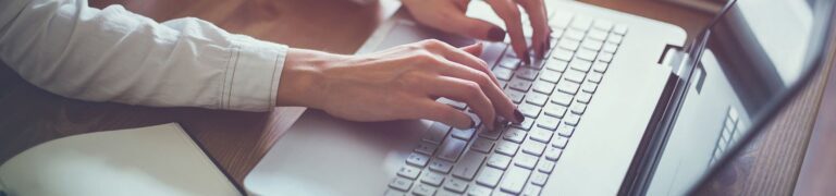 A person wearing a white shirt types on a silver laptop keyboard, with an open notebook and pen nearby on a wooden desk.