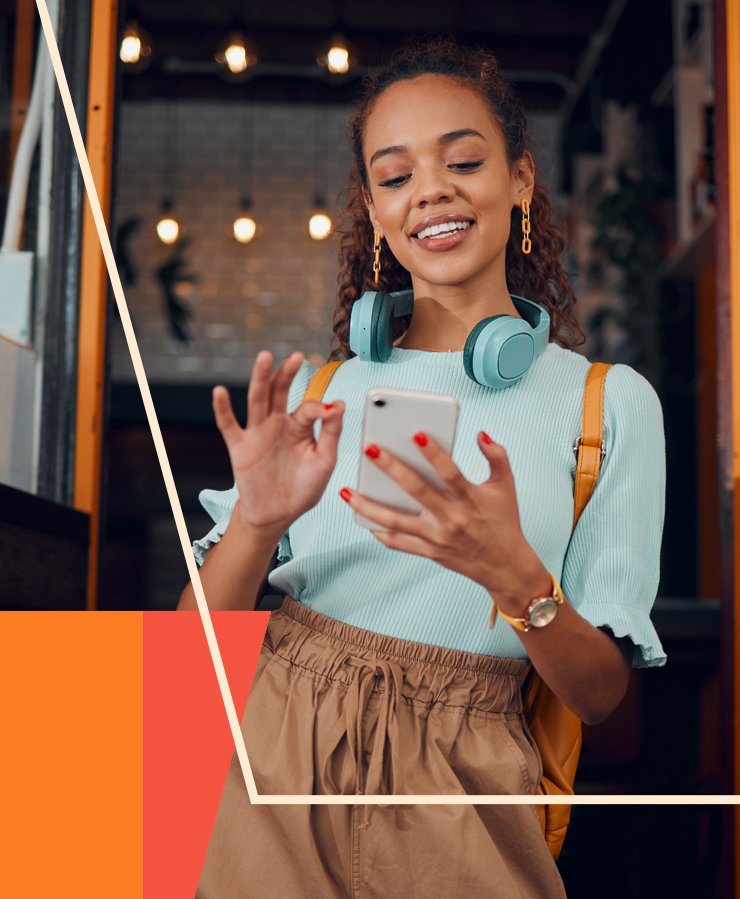 A young woman with headphones around her neck uses a smartphone, smiling and making an OK gesture indoors with warm lighting.