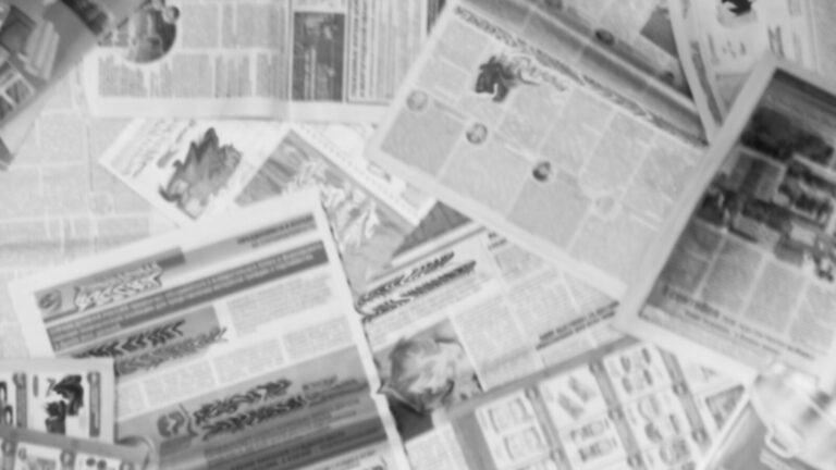 Several newspapers are spread out in an overlapping, scattered manner, with text and images visible but out of focus.