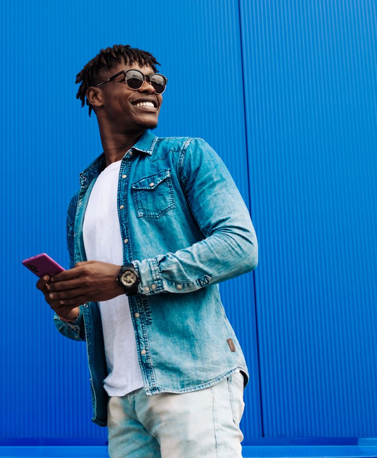 A man wearing sunglasses and a denim shirt over a white t-shirt holds a smartphone and smiles while standing in front of a bright blue corrugated wall.