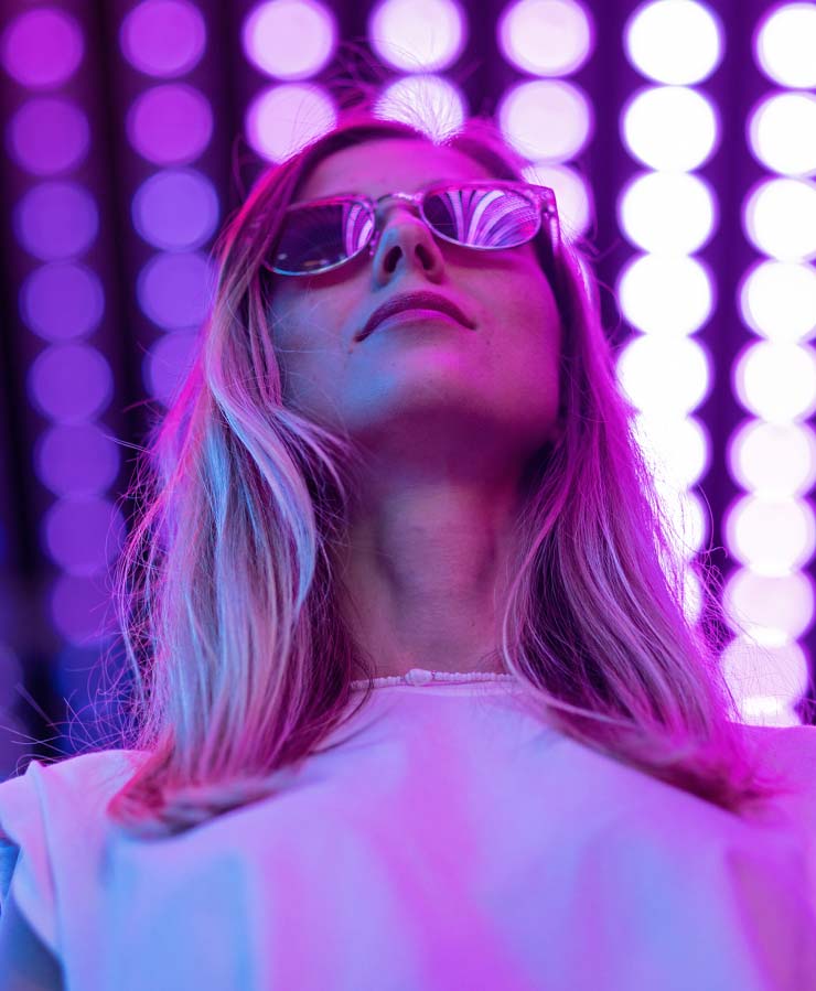 A woman with long hair wearing sunglasses looks upward, illuminated by pink and purple LED lights in the background.