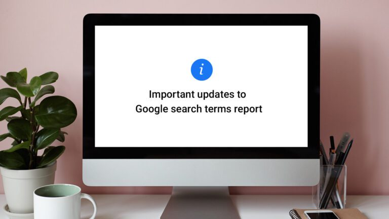 A computer monitor displays a message about important updates to the Google search terms report. A plant, pen, and notebook sit on the desk beside it.