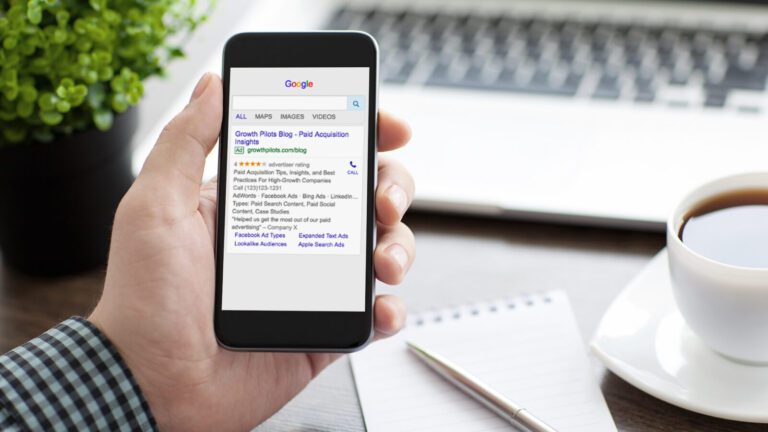A person holds a smartphone displaying Google search results, next to a notepad, pen, and cup of coffee on a desk.