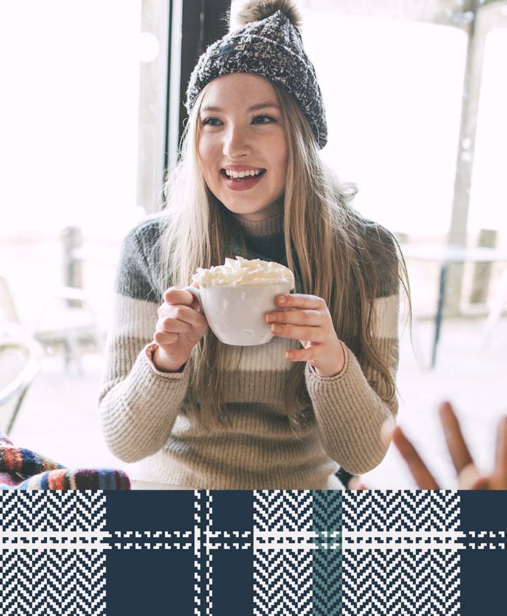 A young woman in a knit hat and sweater smiles while holding a mug topped with whipped cream, seated indoors by a large window.