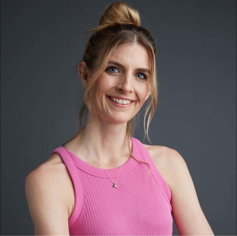 Woman with long blonde hair in a top bun, wearing a sleeveless pink top and necklace, smiling in front of a plain gray background.