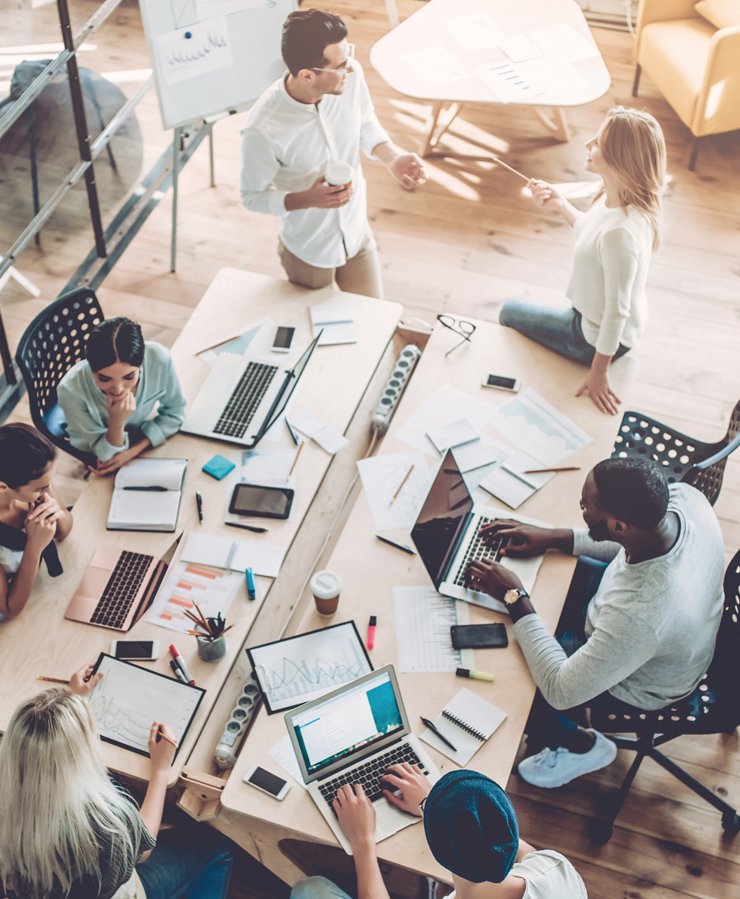 A group of people work at a large table with laptops, papers, and charts; two people stand and converse while others sit and collaborate.