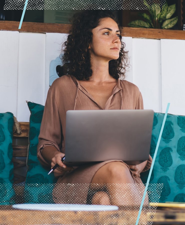 A woman sits with a laptop on her lap and a pen in her hand, looking to the side. She is seated on a couch with teal cushions in a relaxed indoor setting.