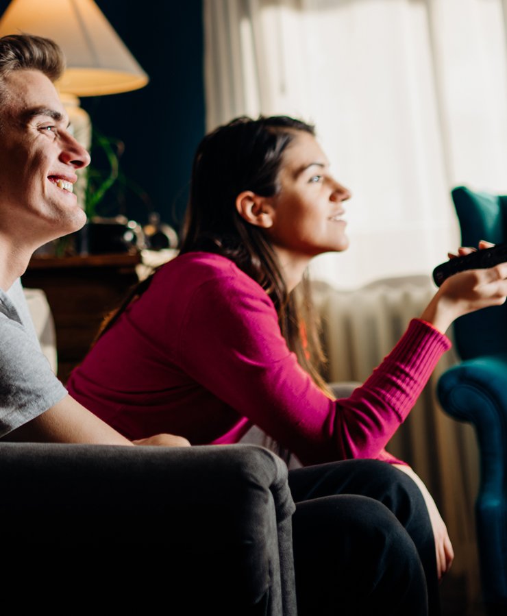 Two people sit on a couch indoors, smiling and watching television. One person holds a remote control.
