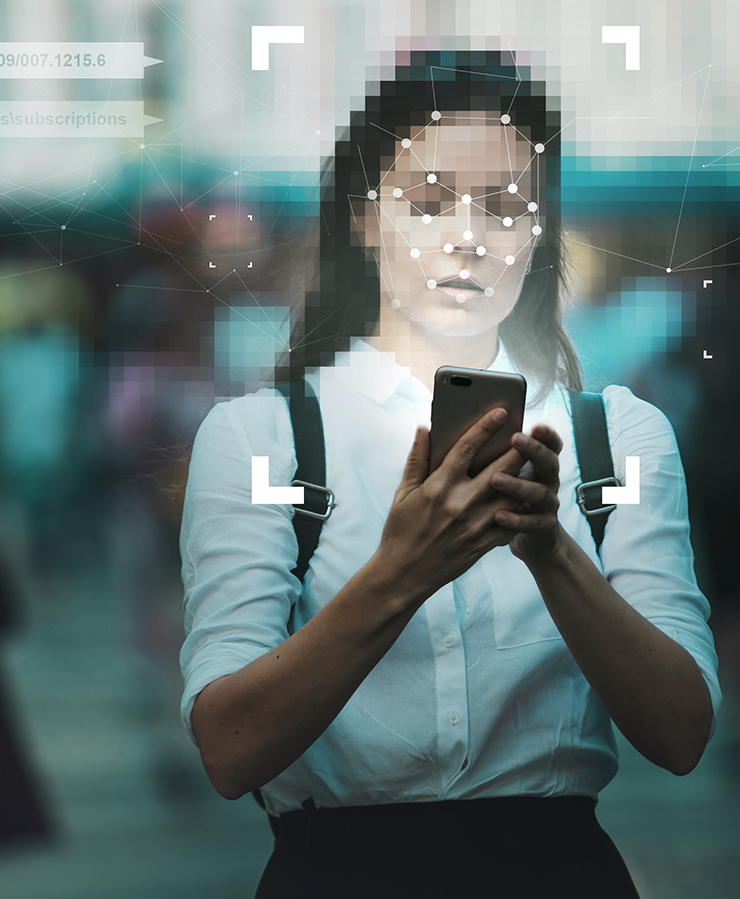 A woman using a smartphone outdoors with facial recognition graphic overlay on her face; blurred background with people.