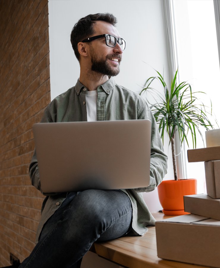 Man sitting by a window with a laptop on his lap, looking outside, with several cardboard boxes and a potted plant nearby.