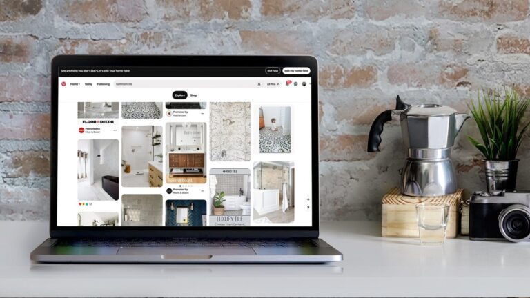 A laptop displaying a Pinterest page with home decor ideas sits on a table next to a moka pot, a camera, and a jar against a brick wall background.
