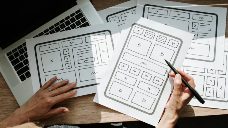 A person reviews website wireframe sketches on paper, with a laptop partially visible underneath the sketches on a wooden desk.