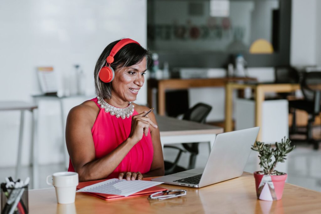 Woman wearing red headphones and a pink top sits at a desk with a laptop, notebook, and coffee cup, smiling and gesturing during an online meeting in a modern office.