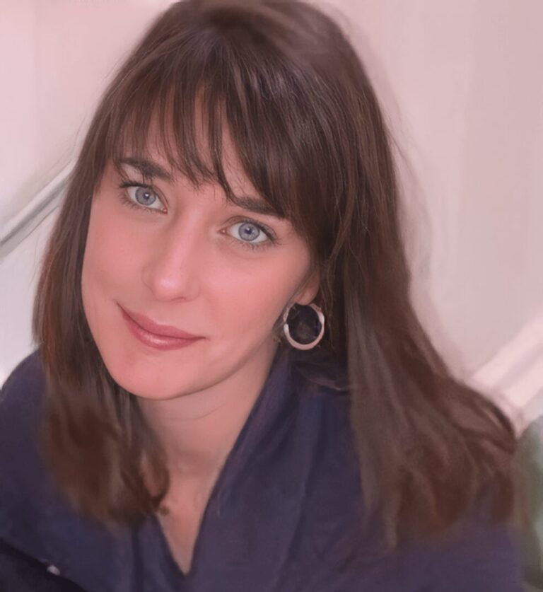 A woman with long brown hair, blue eyes, and hoop earrings looks up at the camera, smiling slightly. She is indoors and wearing a dark top.