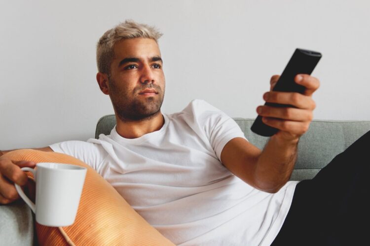 A man with short blond hair sits on a couch holding a TV remote in one hand and a white mug in the other, looking at the television.