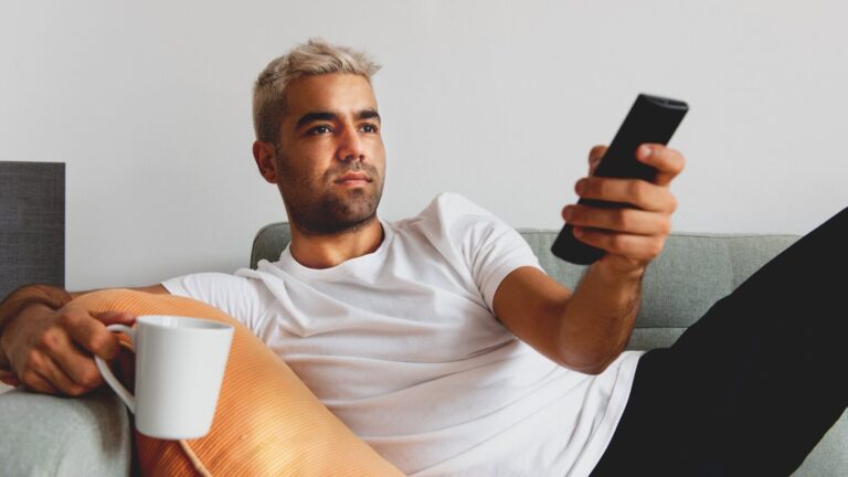 A man with short blond hair sits on a couch holding a TV remote in one hand and a white mug in the other, looking at the television.