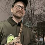 A man wearing glasses, a dark jacket, and a cap stands outdoors at night with a crossbody bag decorated with pins and a plush avocado keychain. Leafless trees and buildings are in the background.