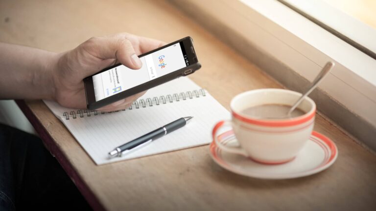 A person holds a smartphone displaying Google search next to a notepad, pen, and a cup of coffee on a wooden table by a window.