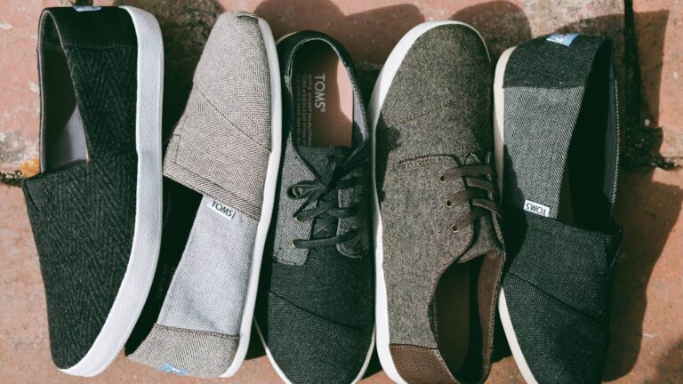 Five pairs of TOMS shoes in various neutral colors and styles are lined up side by side on a stone surface.