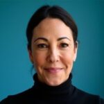 A woman with dark hair pulled back, wearing a black turtleneck, smiles in front of a blue background.